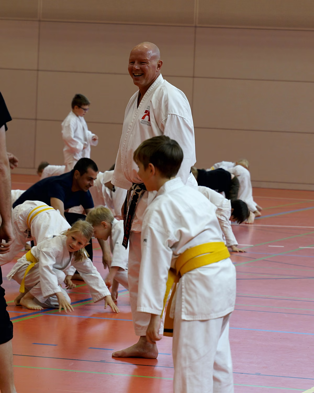 Kinder machen beim Aufw&auml;rmen &Uuml;bungen am Boden, ein Trainer schaut lachend zu