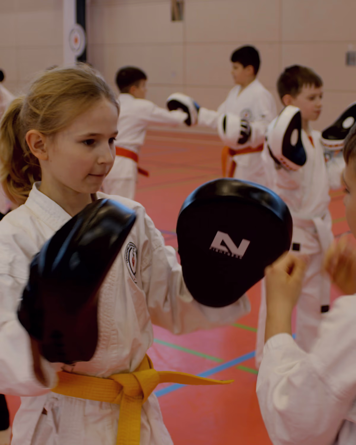 Ein Mädchen im Karateanzug hält beim Kindertraining zwei Pratzen