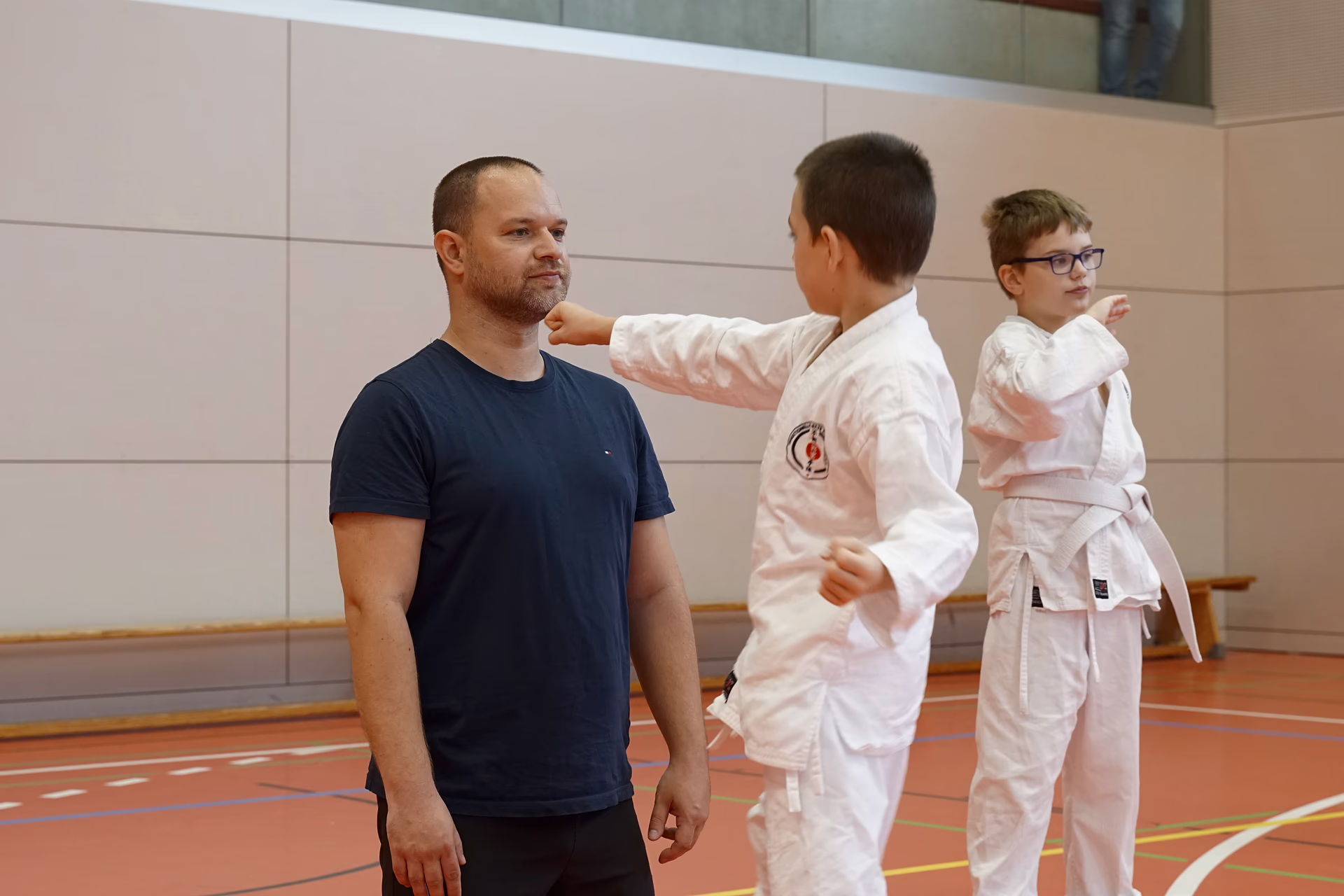 Ein Mann in T-Shirt trainiert mit zwei Jungen im Karateanzug, einer setzt den Fauststo&szlig; an seine Schulter