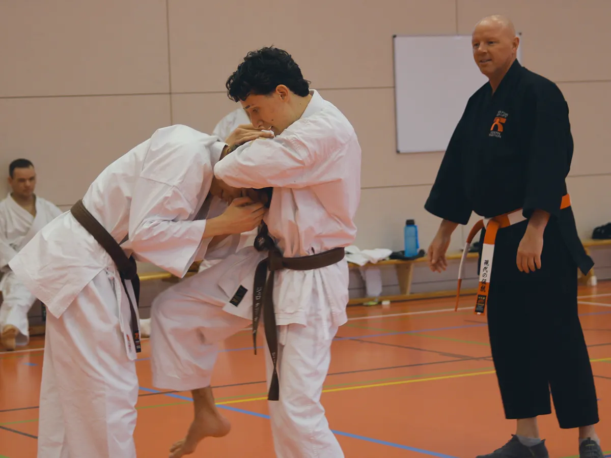 Ein Karateka befreit sich aus einem Schwitzkasten, ein Trainer schaut zu