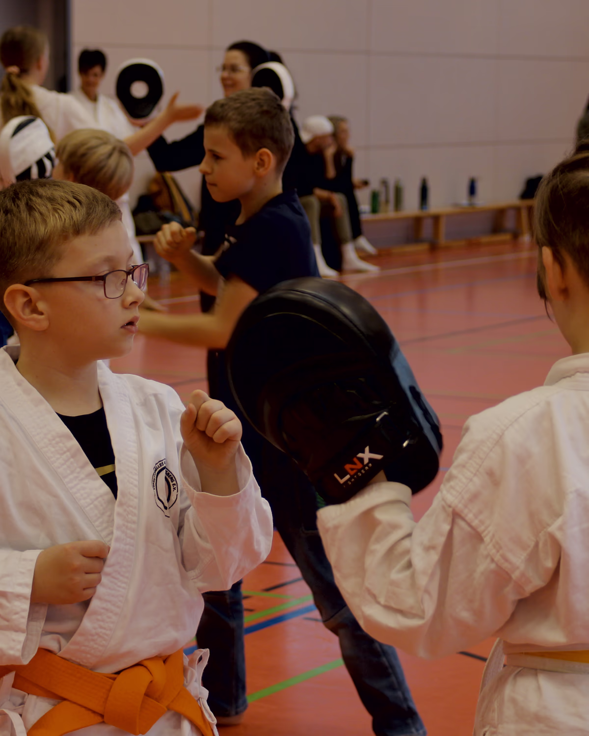 Junge mit Brille und Mädchen trainieren an Pratzen, dahinter weitere Kinderstationen